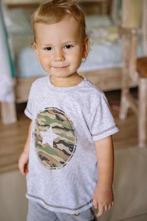 Free Cheerful toddler with a star emblem shirt standing indoors on a wooden floor. Stock Photo