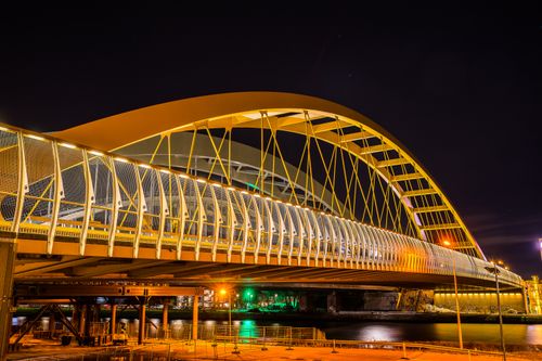 Free A beautifully lit modern arch bridge spans a river, glowing against a night sky. Stock Photo