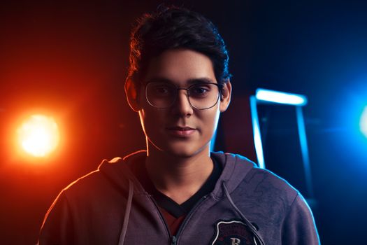 A young man wearing glasses is dramatically lit by vibrant colored lights, creating an intense mood.