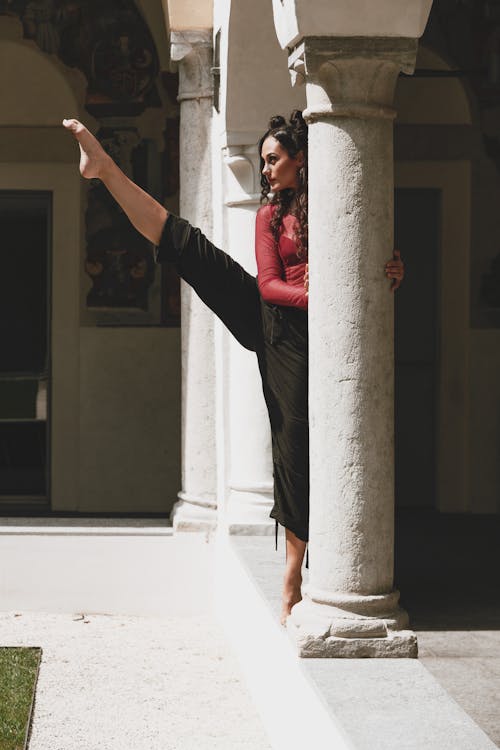 Full body of serious graceful female dancer with leg raised standing near stone column