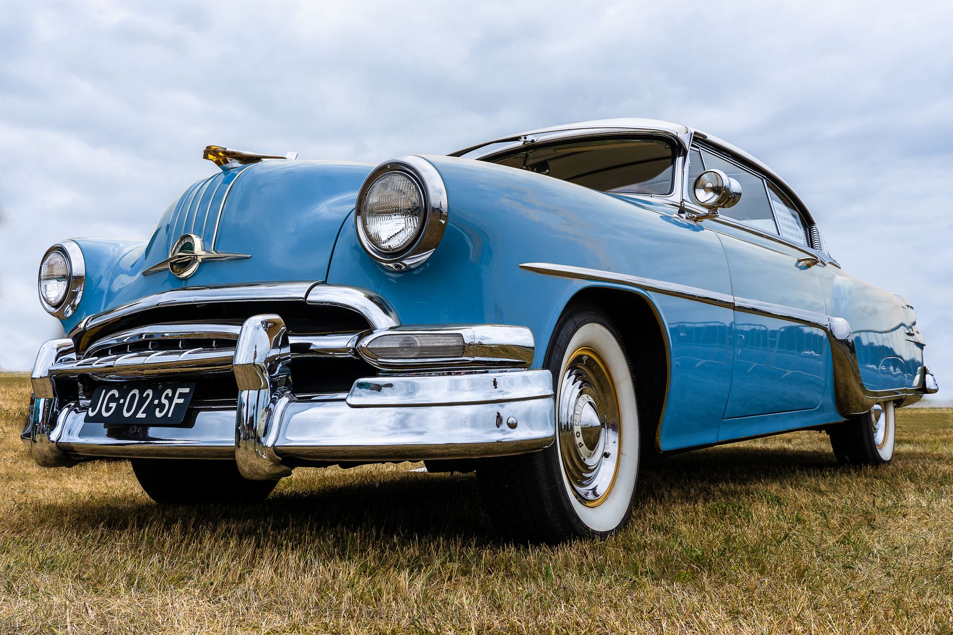 A beautifully restored blue vintage car with chrome details parked on a grass field under a cloudy sky.