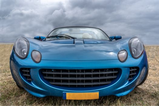 Front view of a classic blue sports car in an open field under a dramatic cloudy sky.