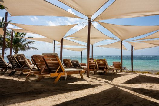 A serene beach scene featuring sun loungers under stylish shade sails by clear blue water.