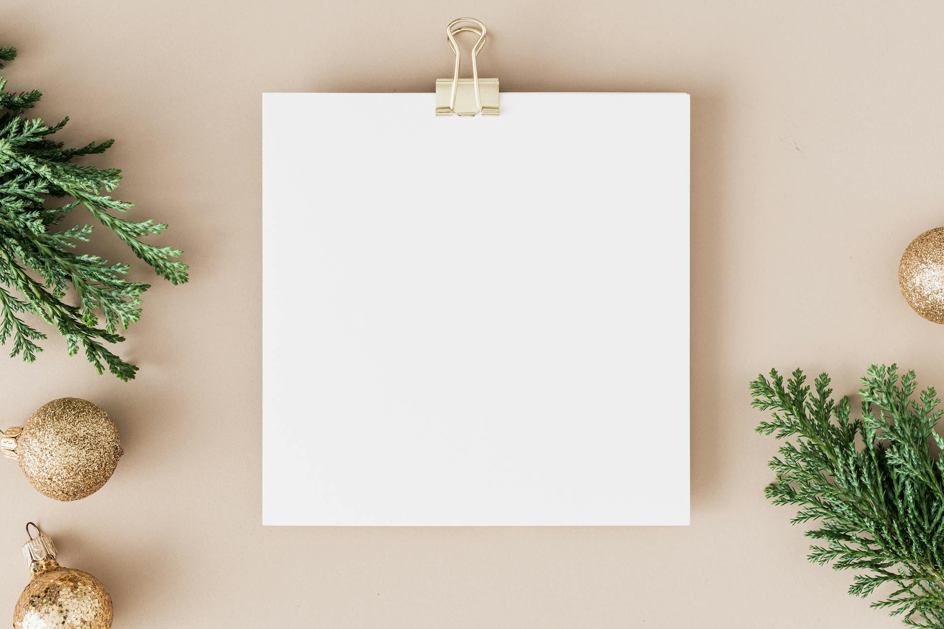 Christmas mockup featuring blank paper clip bordered by gold ornaments and pine on beige background.