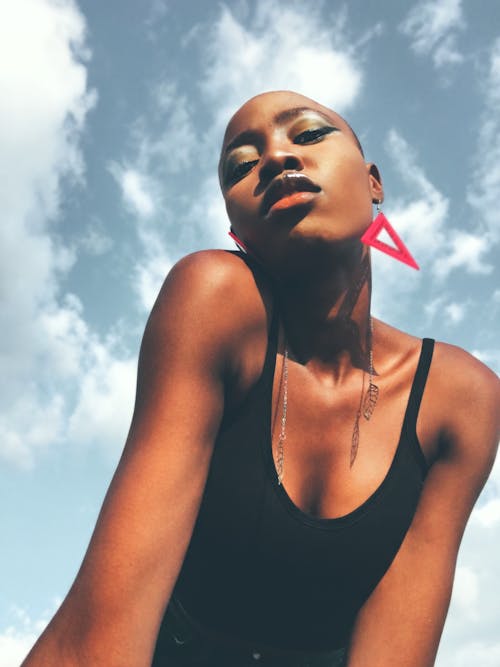 From below of young stylish confident black lady with short hair in black top and accessories leaning forward and looking at camera against blue sky