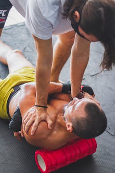 A trainer helps an athlete stretch using a foam roller for muscle relaxation indoors.