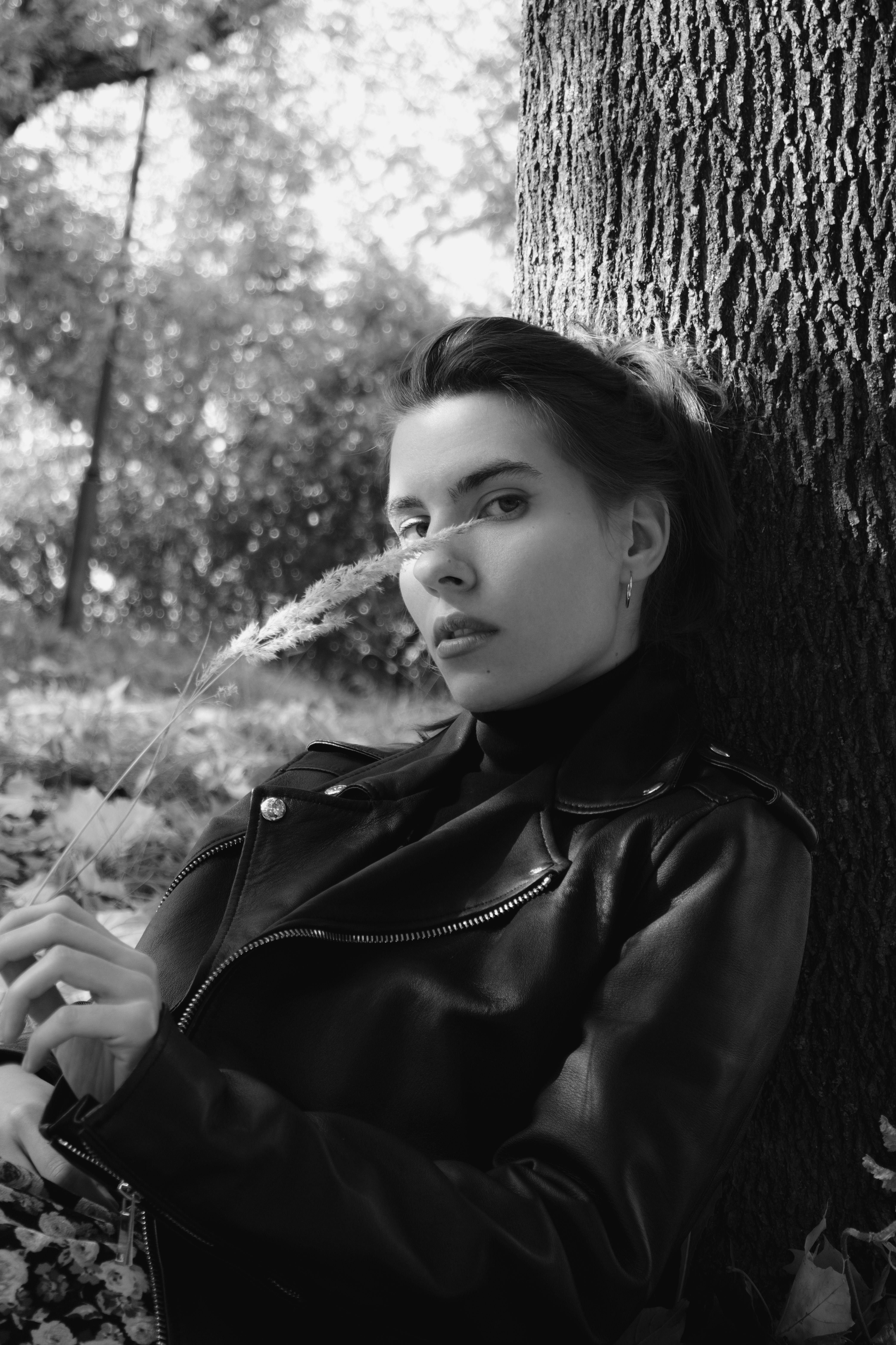Artistic black and white portrait of a woman in a leather jacket leaning against a tree outdoors.