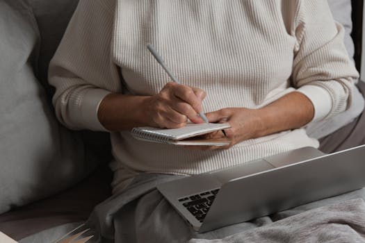 A person writing in a notebook with a pen while sitting by a laptop, creating a cozy work atmosphere.