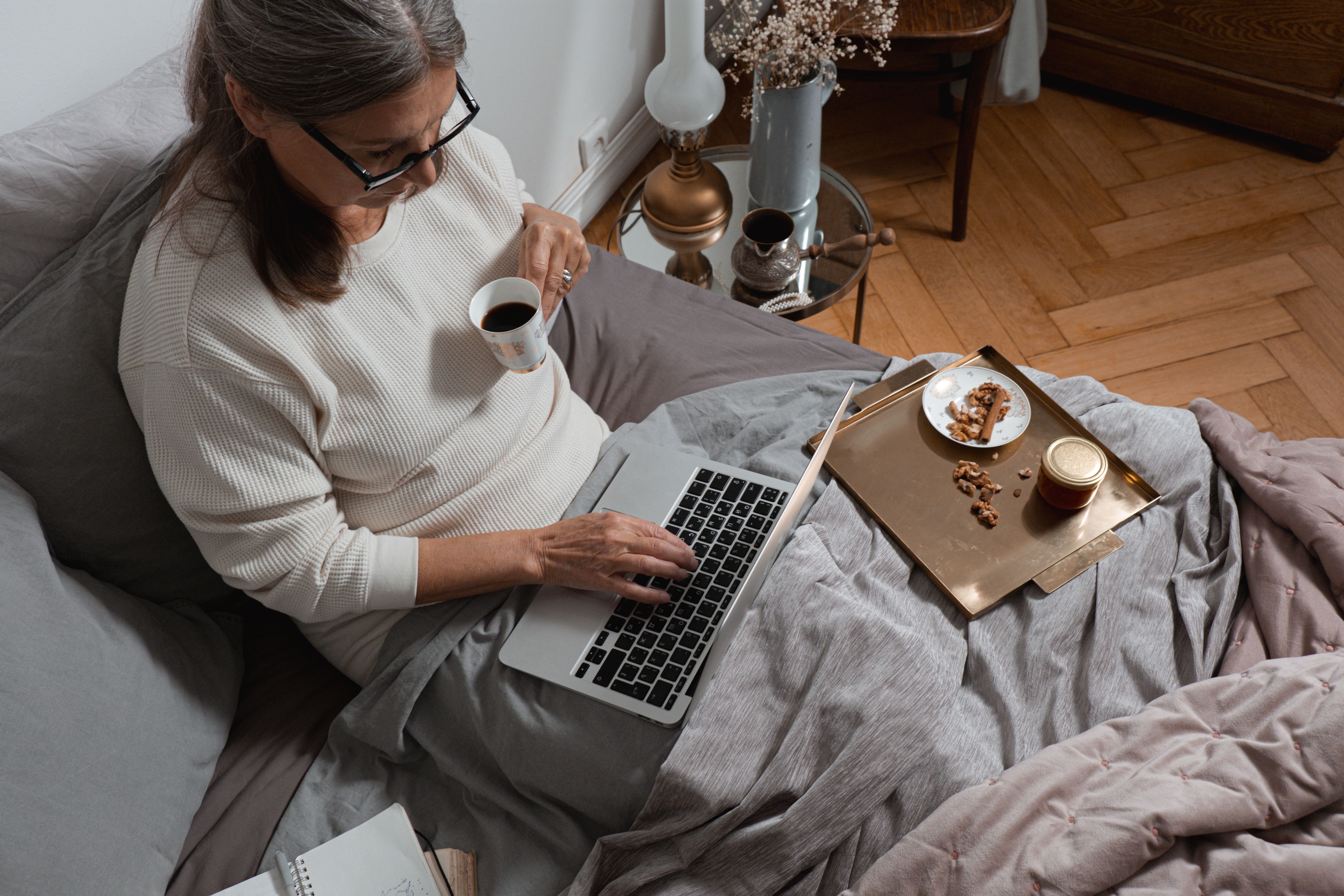 Senior woman comfortably working from bed with coffee and snacks, showcasing remote work lifestyle.