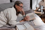 一位年长的女性戴着眼镜，铺着舒适的毯子，躺在床上舒适地读书，享受着宁静的时刻