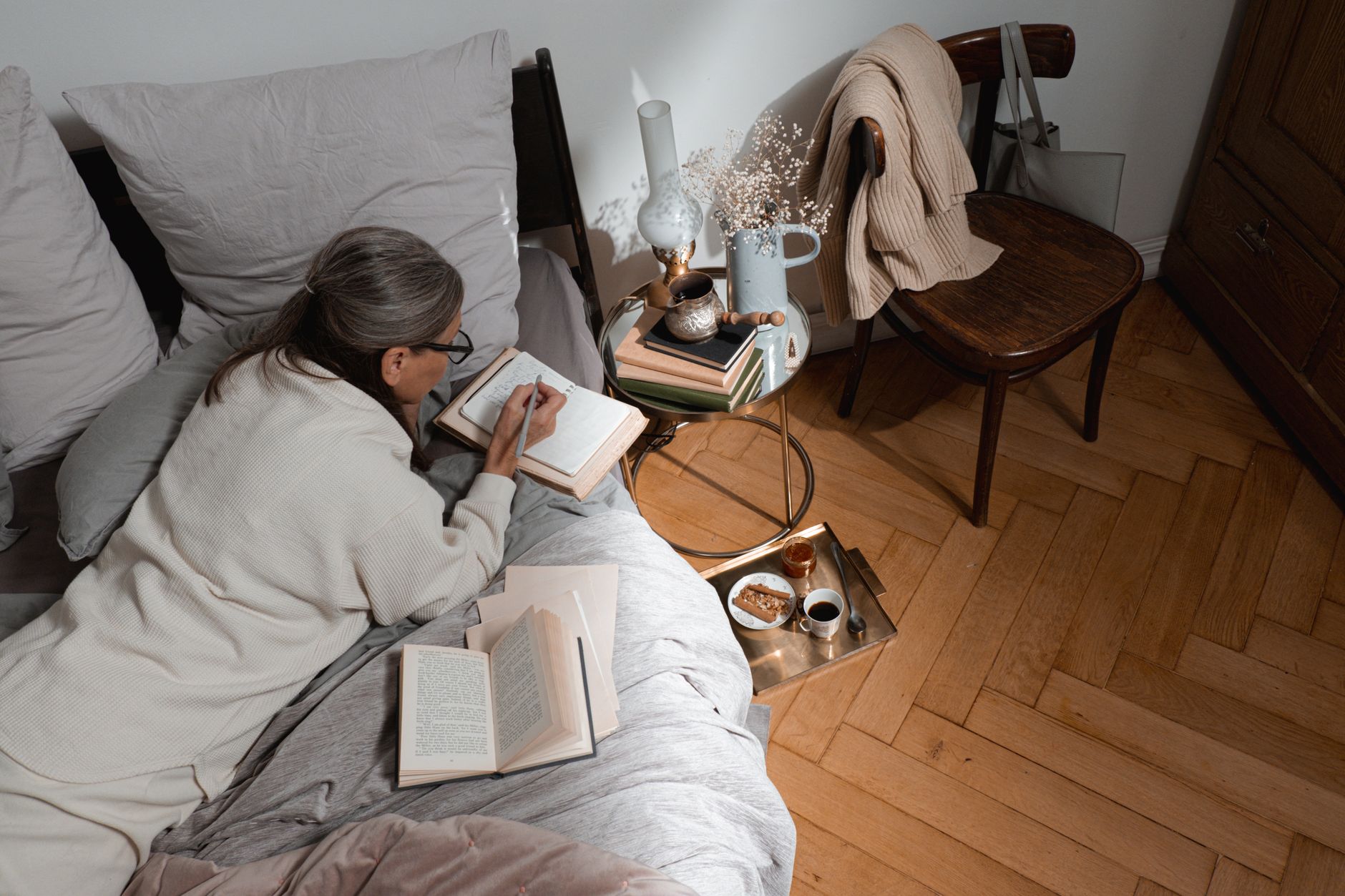 Senior woman takes notes in bed with coffee, books, and cozy decor on a peaceful morning.