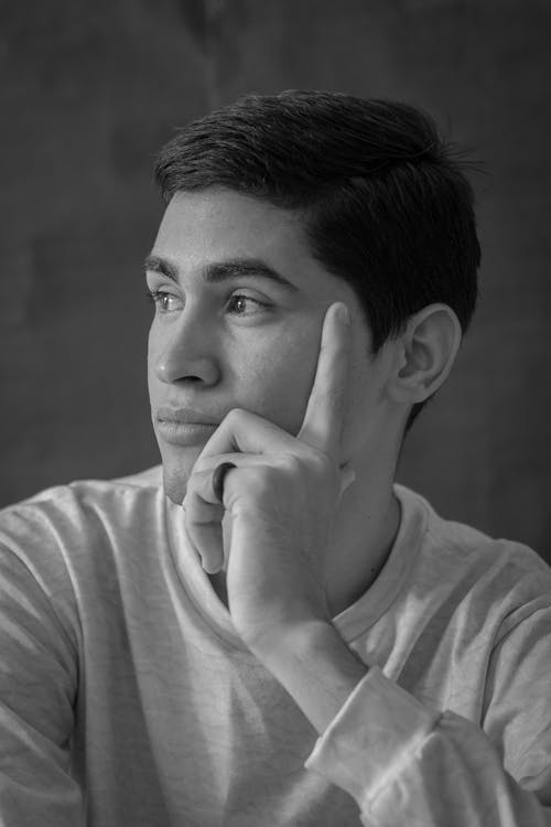 Black and white portrait of handsome young male in casual clothes touching cheek and looking away in contemplation