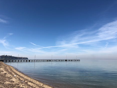 A tranquil beach scene with a long pier extending into a calm ocean under a clear blue sky.