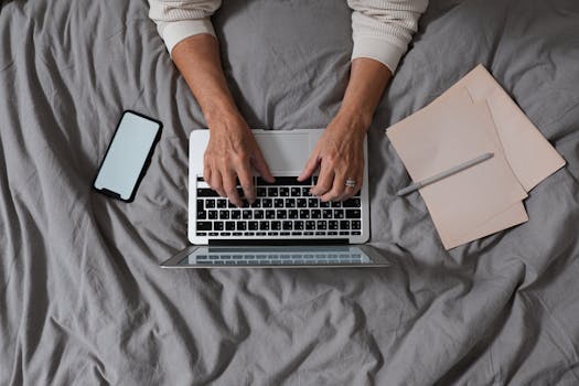 Hands typing on a laptop with smartphone and papers on bed. Cozy workspace.