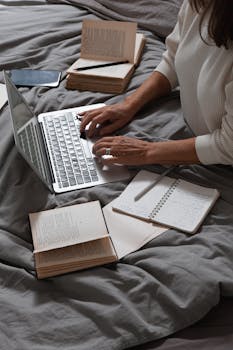 A woman types on a laptop surrounded by books and a notebook on a bed, suggesting a cozy work environment.