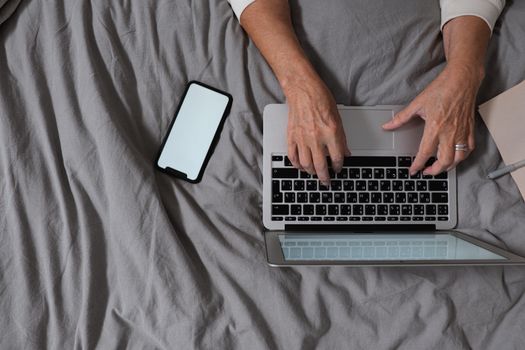An adult typing on a laptop with a smartphone beside her on a grey bed, depicting a home office setting.