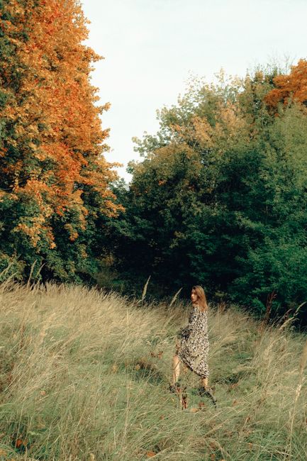 Side view of anonymous female in trendy outfit strolling on grass meadow near autumn trees in countryside