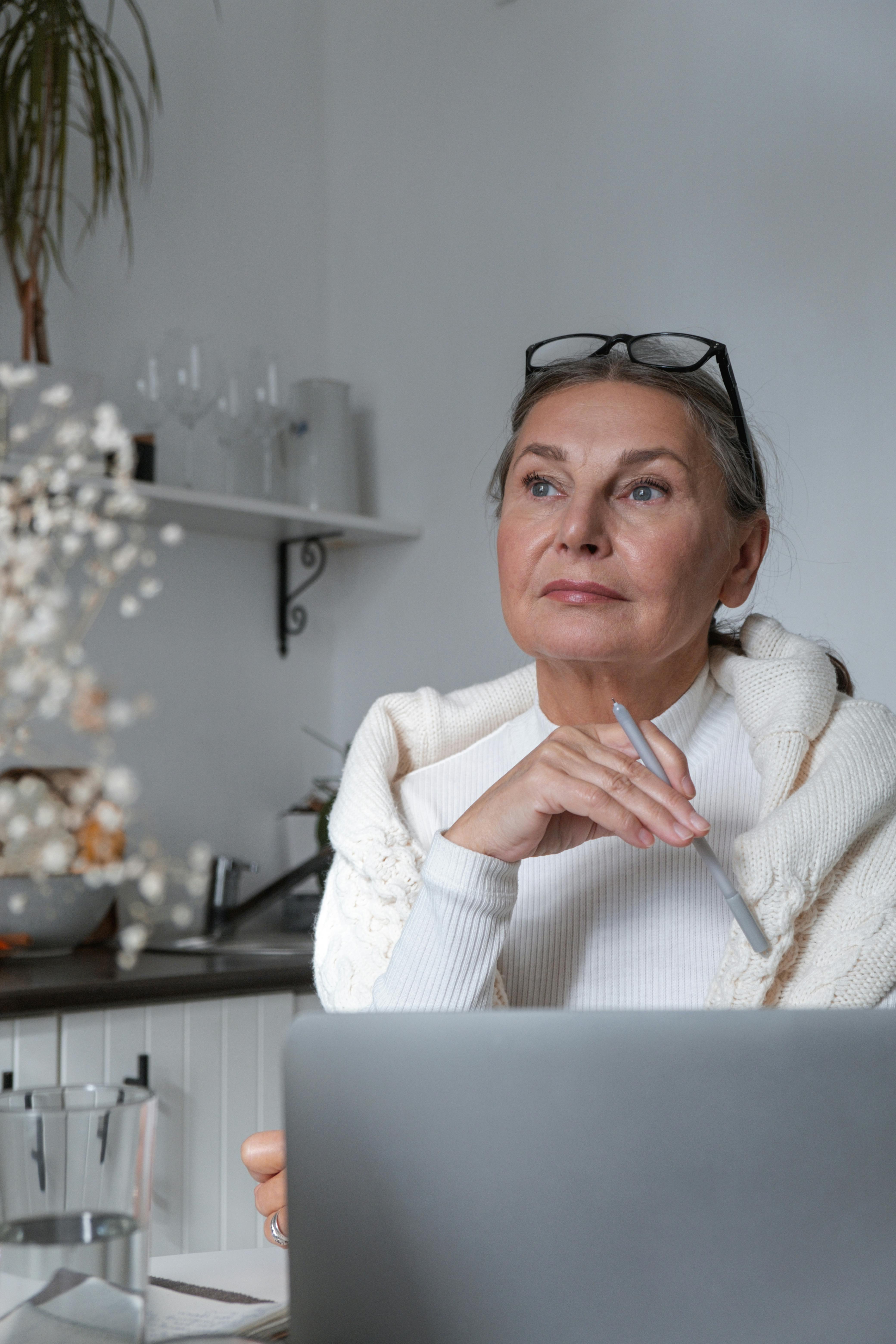 一位资深女性戴着眼镜，在舒适的室内环境中若有所思地使用着笔记本电脑