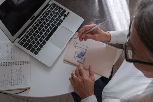An adult woman sketching in a notebook at a desk with her laptop, working from home.