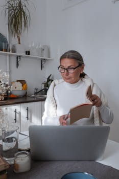 Elderly woman in glasses reading a book while working on a laptop in her cozy kitchen.