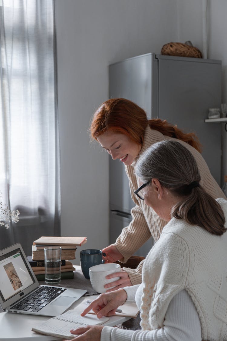 Two Women Using A Laptop In A Kitchen And Drinking Coffee 