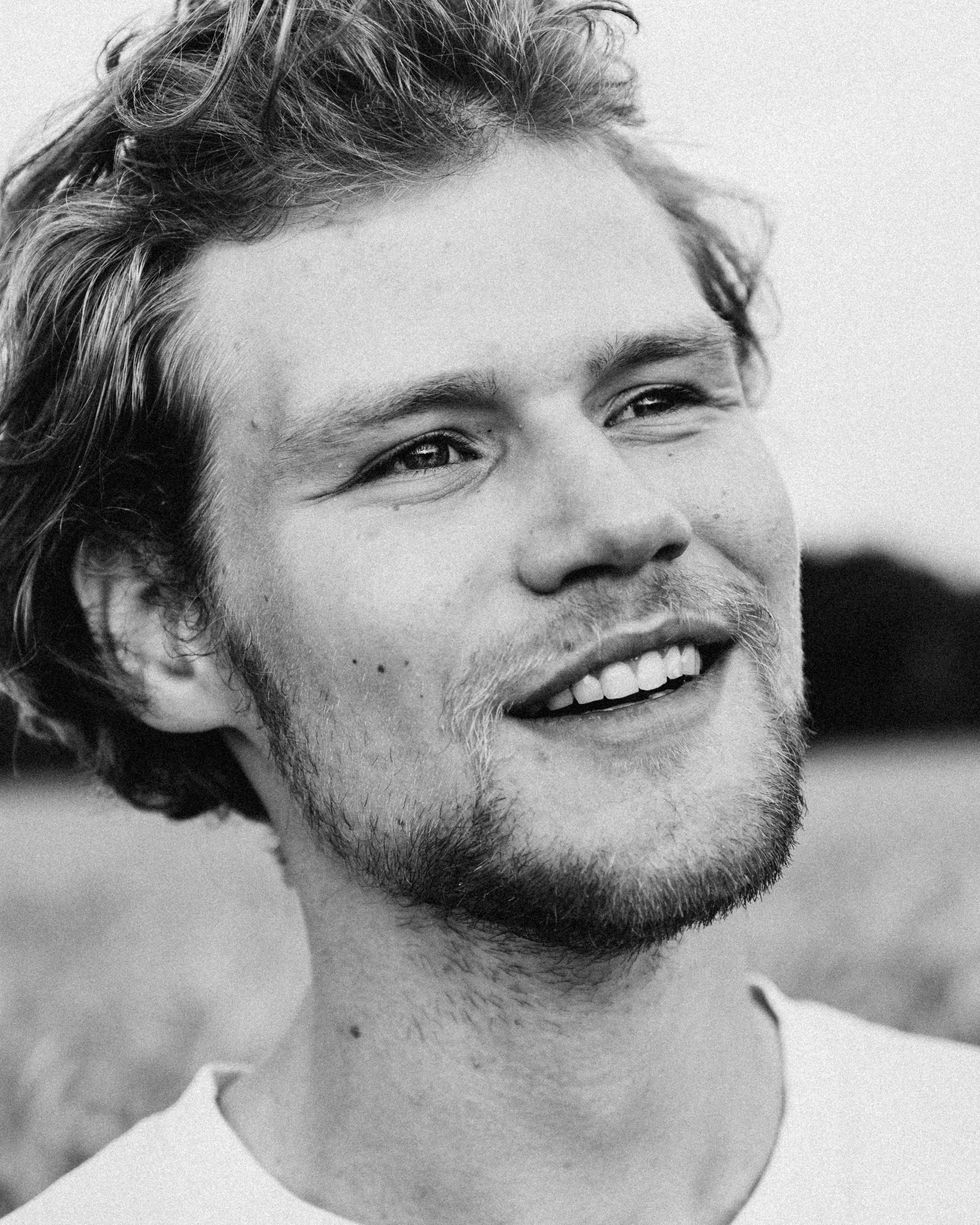 Black and white close-up portrait of a smiling young man with tousled hair and a beard, looking away from the camera in a field