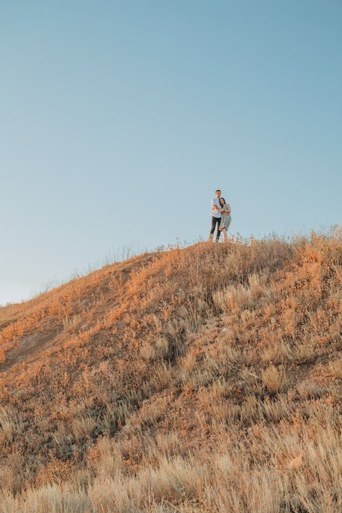 Unrecognizable distant romantic couple hugging while standing on top of mount with dry grass against blue sky in sunny day