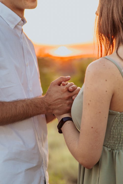 Side view of crop anonymous young man holding hand of girlfriend during date in countryside against sunset sky