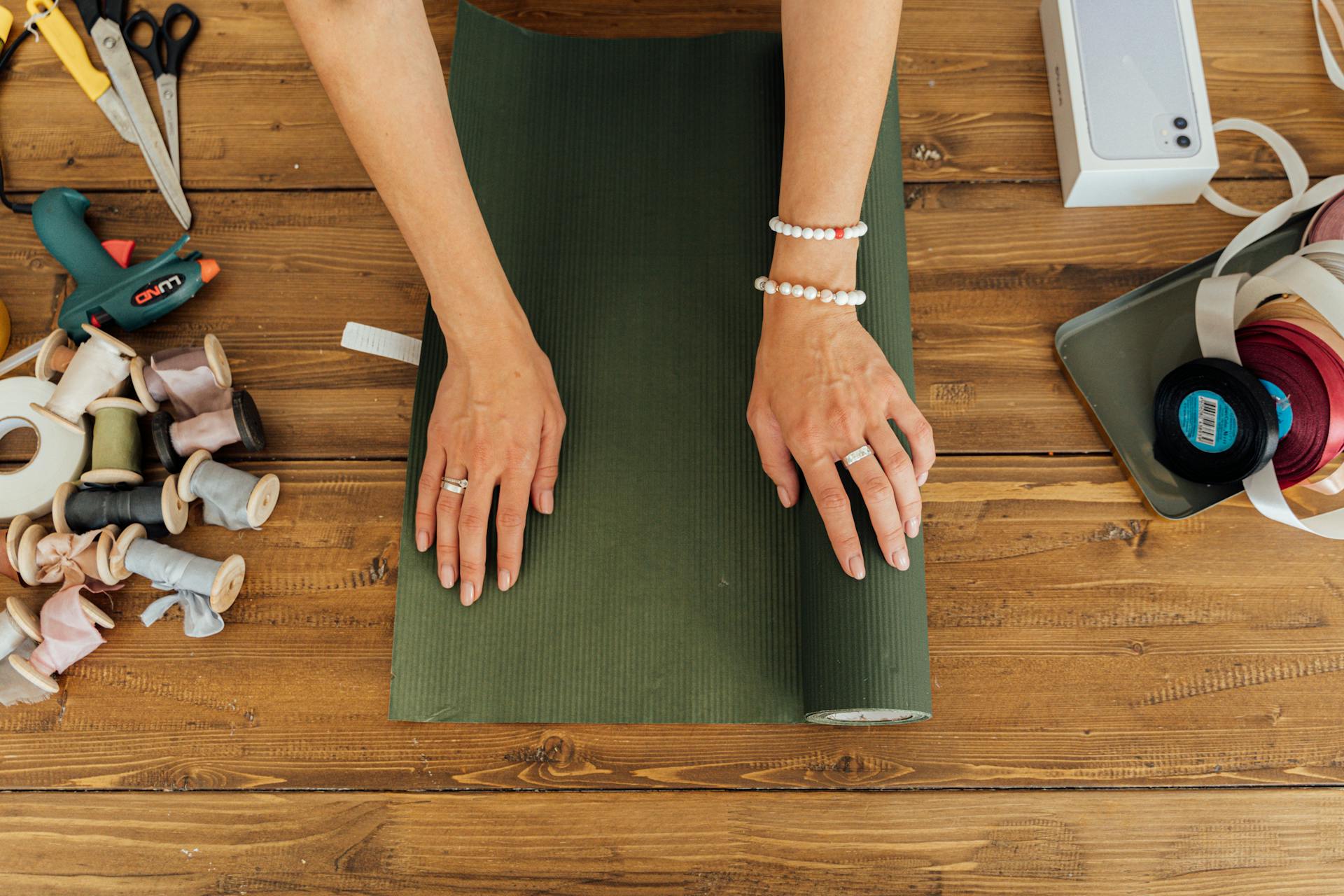 Hands wrapping a gift with green paper, surrounded by crafting tools and materials, ideal for DIY projects.