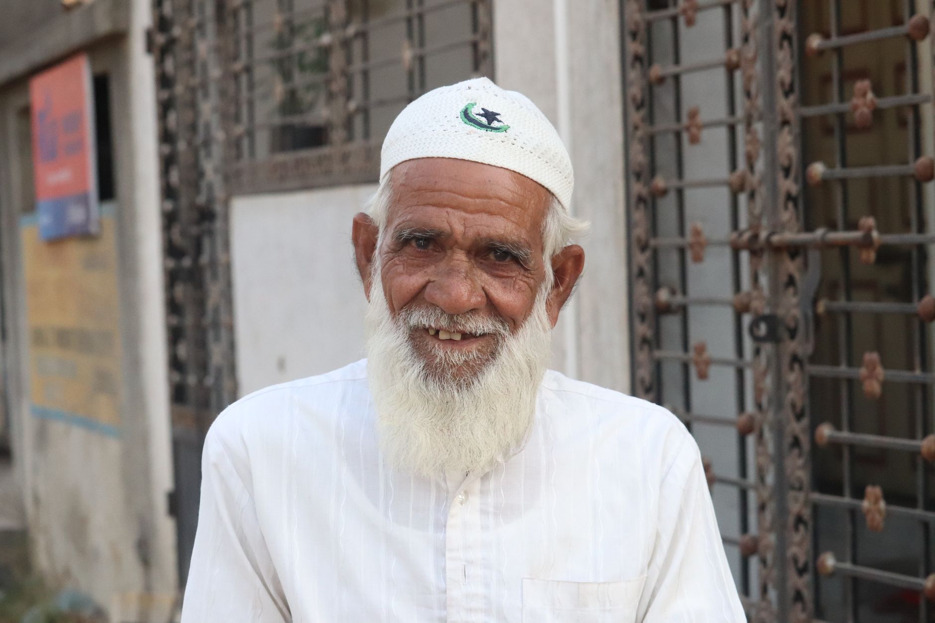 Portrait of an elderly man with a white beard wearing traditional attire and a skull cap, smiling warmly.