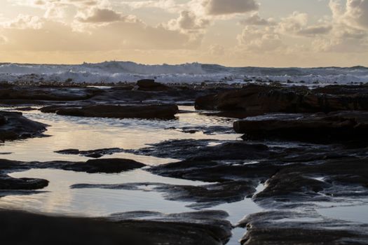 Capture the tranquility of a rocky coastline in Cape Town during sunset with waves crashing ashore.