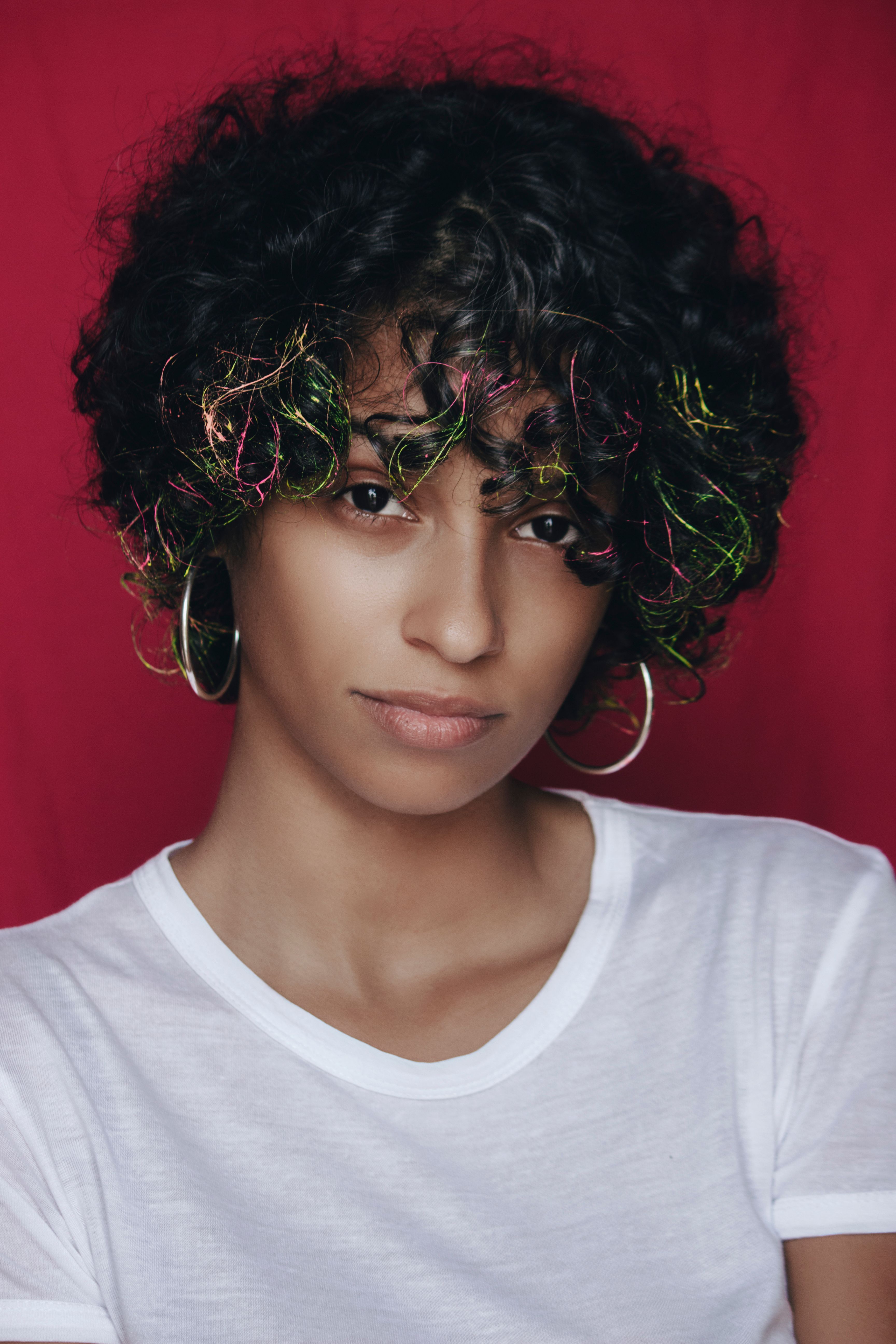 Close-up portrait of a woman with curly hair, colorful highlights, and hoop earrings against a red background.