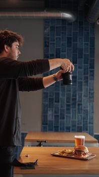 A photographer takes a vertical shot of a burger and beer on a wooden table indoors.