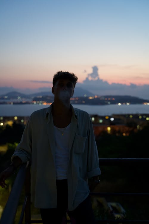 Young man in casual outfit on balcony of high building looking at camera and exhaling smoke in evening