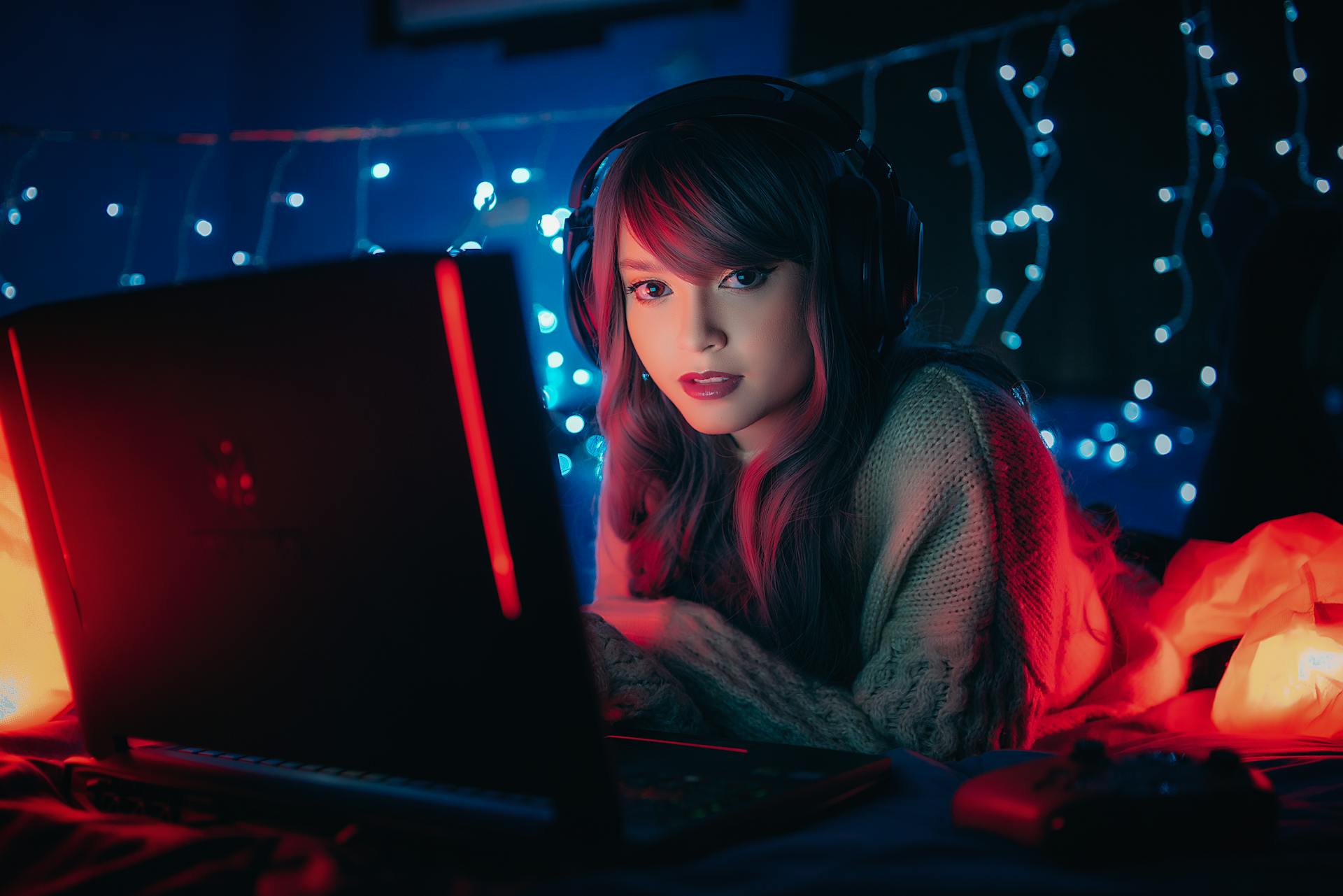 Young woman wearing headphones, gaming on a laptop surrounded by dreamy string lights. Cozy ambiance.