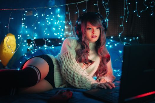 Young woman in cozy sweater gaming at night with glowing Christmas lights, creating an atmospheric ambiance.