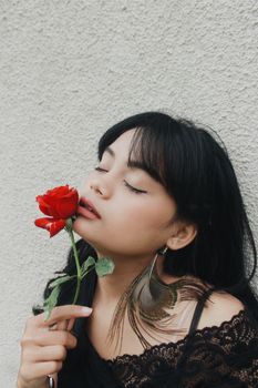 一位迷人的年轻少数族裔女性，闭着眼睛靠在白色灰泥墙上，闻着新鲜红玫瑰的香气，她留着长长的黑发