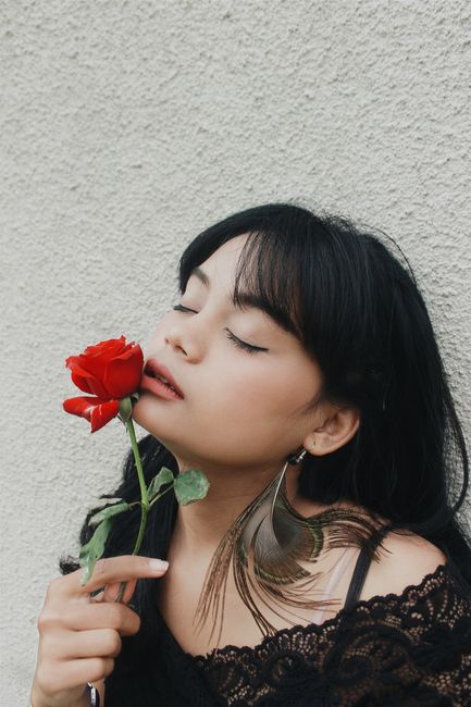 Alluring young ethnic female with long dark hair enjoying smell of fresh red rose with closes eyes against white stucco wall
