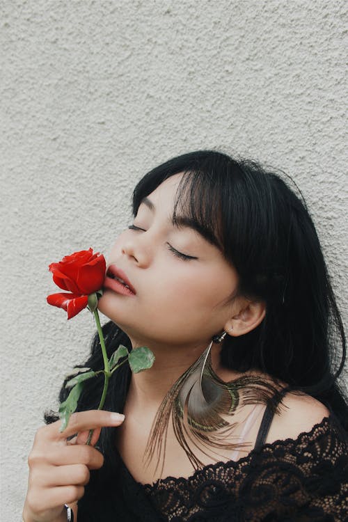 Free Alluring young ethnic female with long dark hair enjoying smell of fresh red rose with closes eyes against white stucco wall Stock Photo