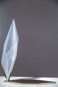 A minimalist vertical glass sculpture displayed on a sleek metal stand with a gray background.