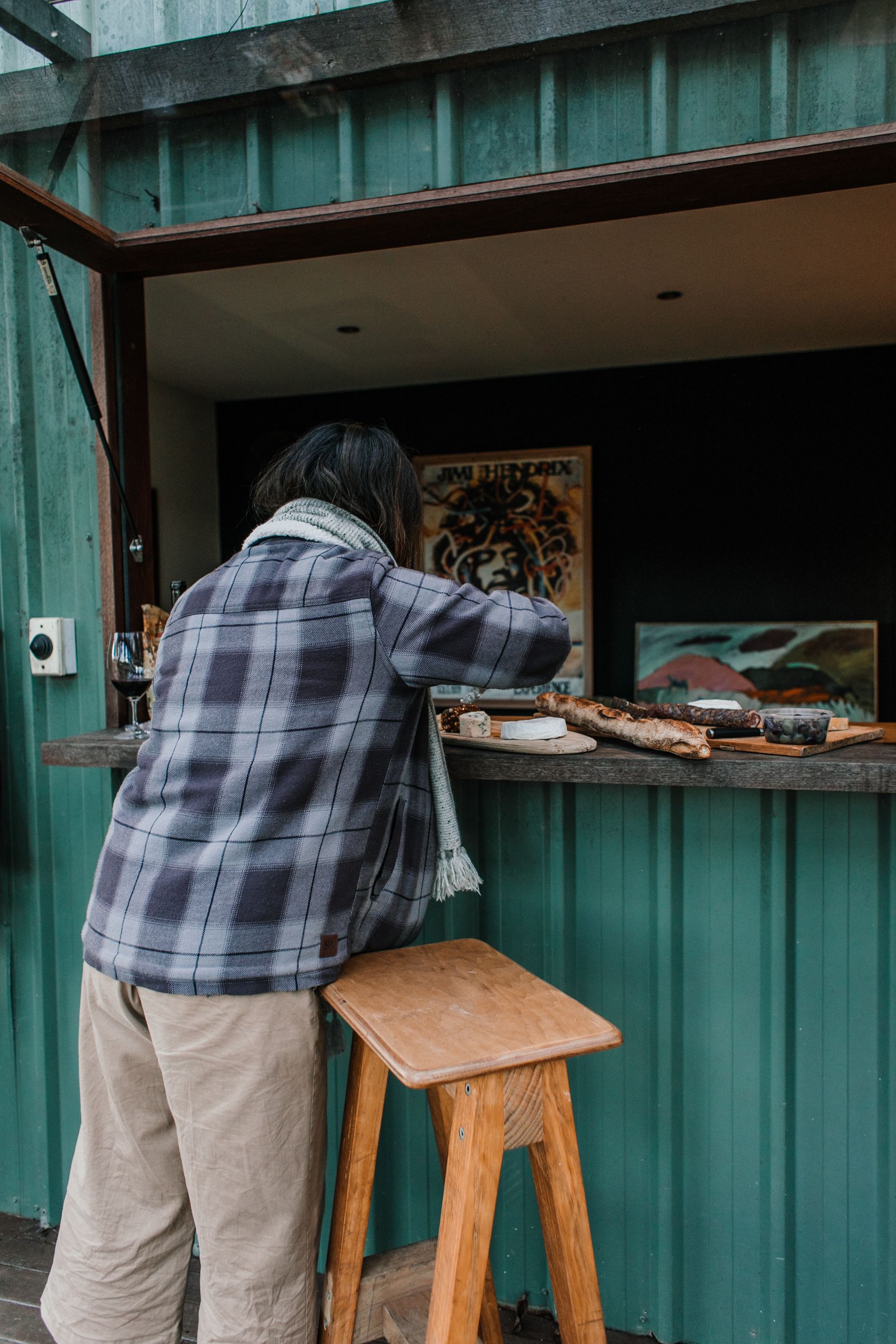 Back view of anonymous person cutting fresh cheese on wooden bard while standing at green stall near chair on street