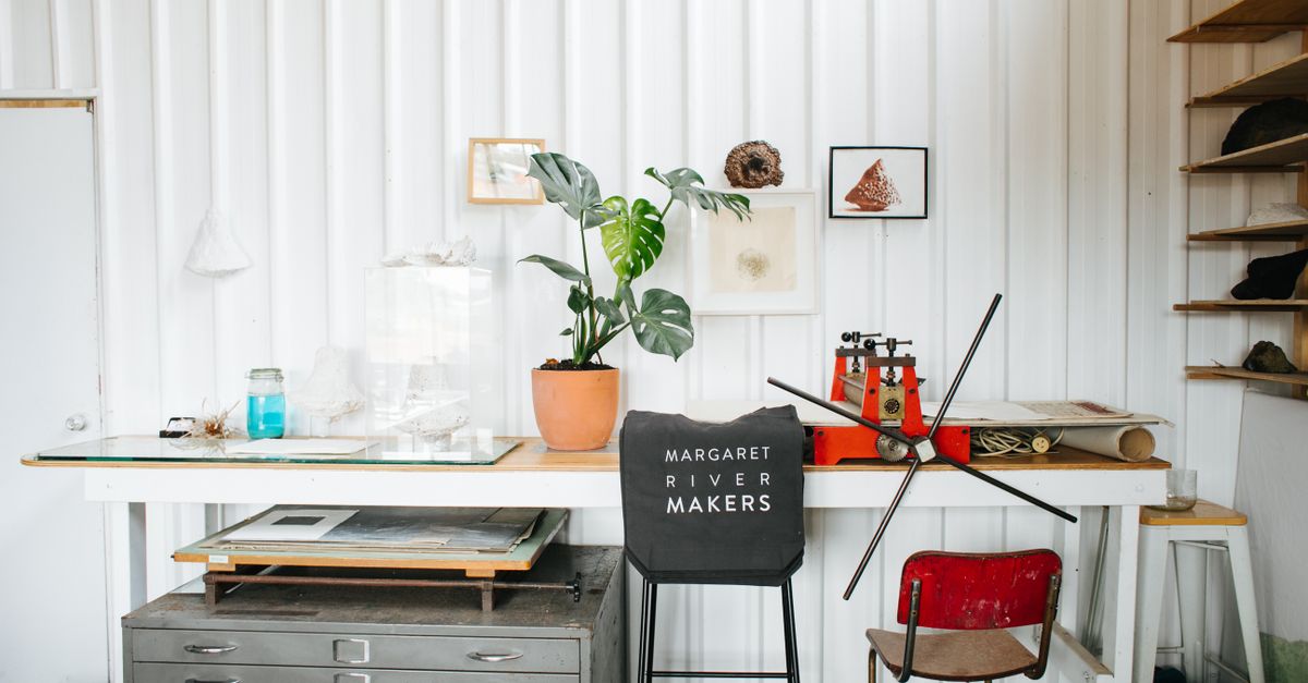 Interior of creative workplace with printing press and flowerpot placed on table near drawer and chair with manufactured product with inscription