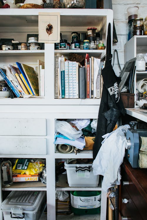 White cupboard with collection of books and abundance of assorted materials and supplies placed on shelves with containers in professional studio