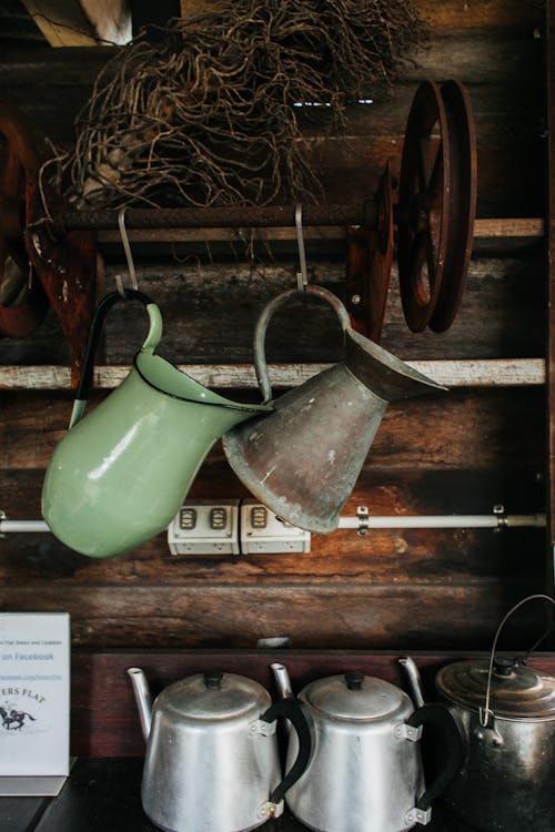 Retro metal jugs hanging on hooks at shabby wooden wall near old kettles and pot in old fashioned rustic cottage