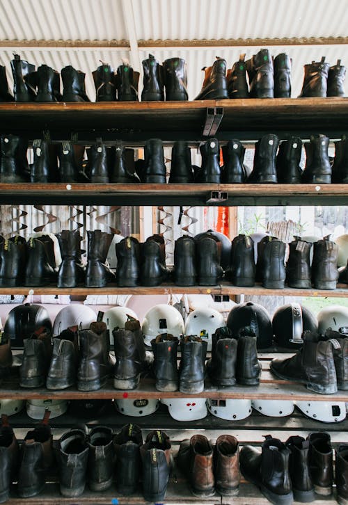 Abundance of leather old fashioned boots placed on wooden shelves near plastic protective helmets for rent on street in city