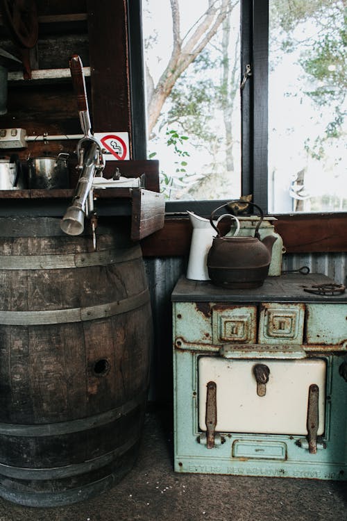 Aged kettle and dishes placed on old cooker near weathered barrel at window in vintage styled room in suburb area