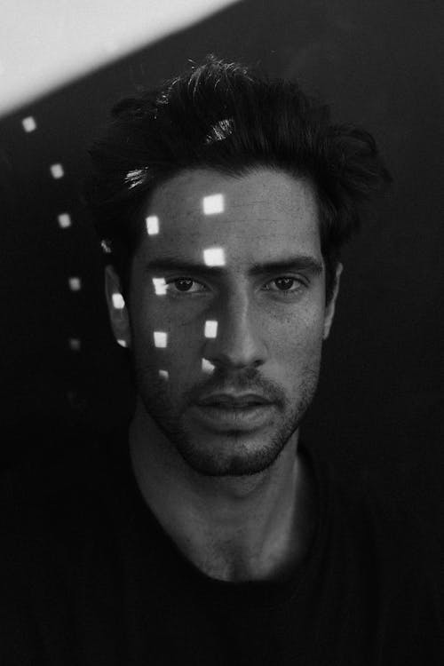 Black and white of confident bearded male looking at camera while standing under shadow with bright light glares on face