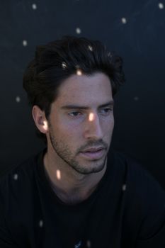 从上方看，一位留着胡须的梦幻男性站在黑色背景上，脸上映着强光，目光凝视着别处