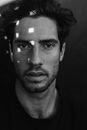 Black and white of bearded male with dark hair looking at camera while standing under shadow with sunlight on face