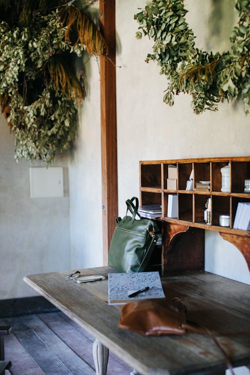 Aged room with shoulder bag near book with pen on wooden table under plant wreaths in building
